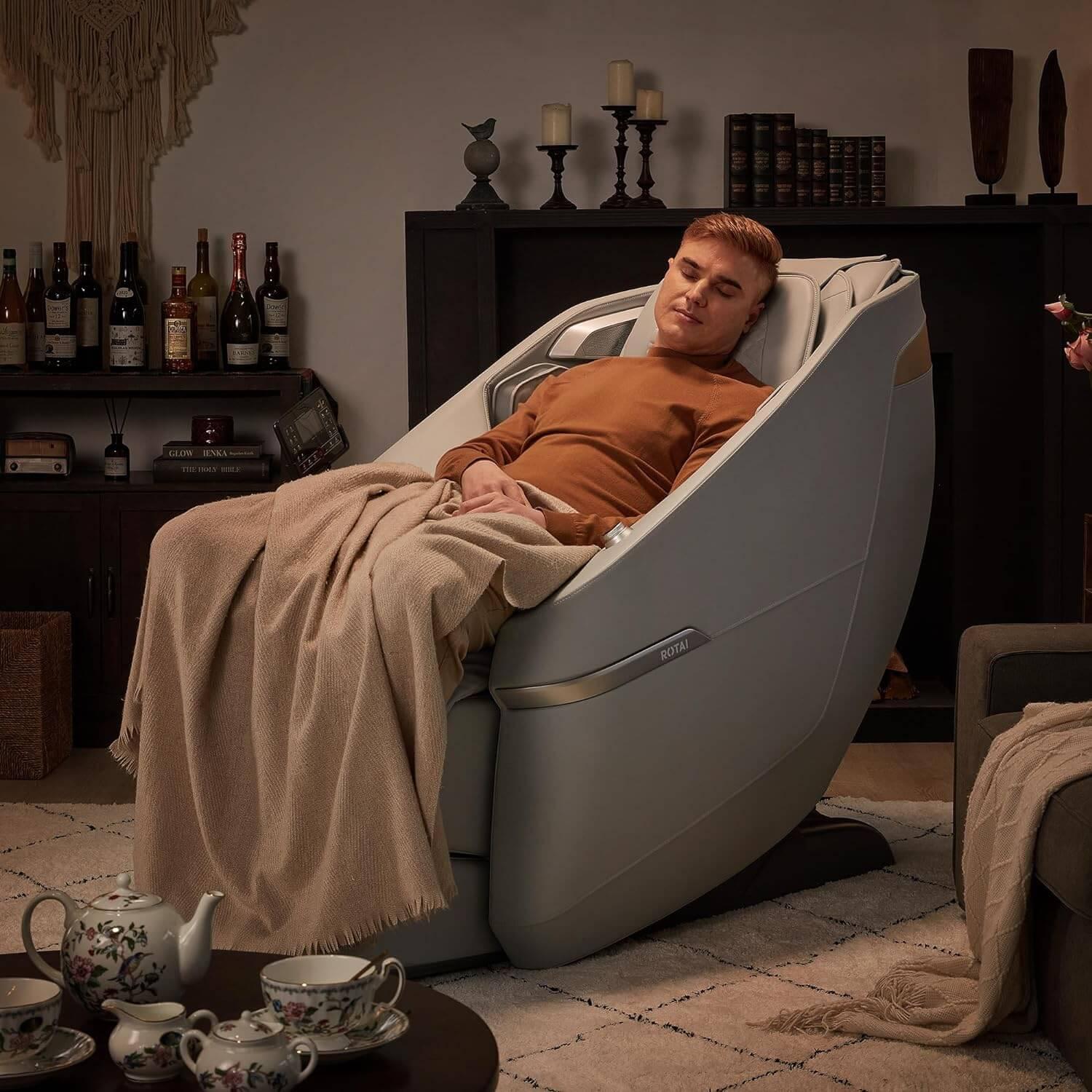 Man relaxing in Ekanite massage chair and sofa in cozy living room with a blanket, showcasing tablet control and foot massager features.